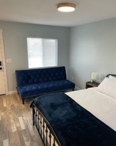 Home bedroom interior. Blue velvet sofa, bed with white linens, wood-look flooring, natural light.