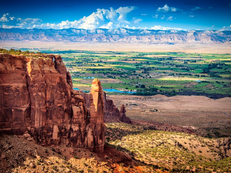 Red rock cliffs overlook a lush green valley with scattered farms, while distant mountains rise under a blue sky. This stunning scene shows why october-is-a-great-time-to-visit-palisade, blending desert views with greenery and rugged peaks.