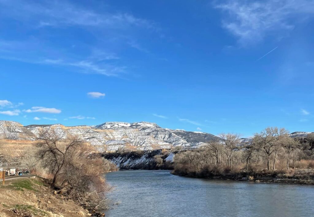 A wide river curves through the Grand Valley with leafless trees along its banks. In the distance, snow-capped mountains rise under a bright blue sky with a few wispy clouds.