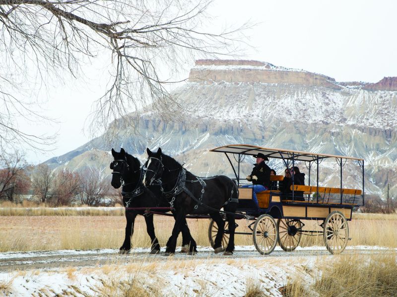 A horse-drawn carriage with two black horses travels along a dirt path in a snowy, grassy landscape with rocky mesas and leafless trees in the background.