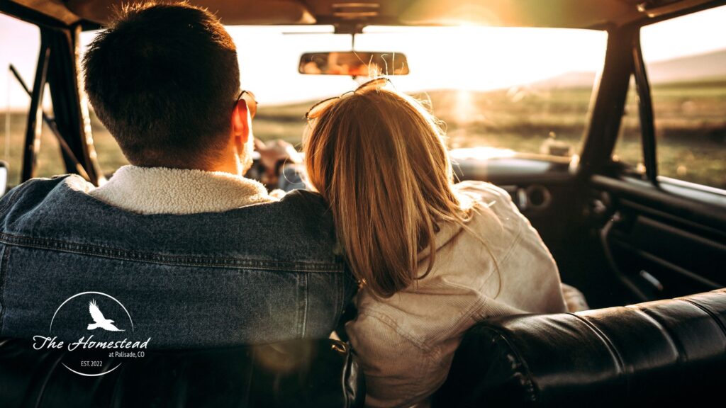 A couple sits closely together in the front seat of a car, looking out at a bright sunset over open fields. The woman leans her head on the mans shoulder. The Homestead logo is visible in the lower left corner.