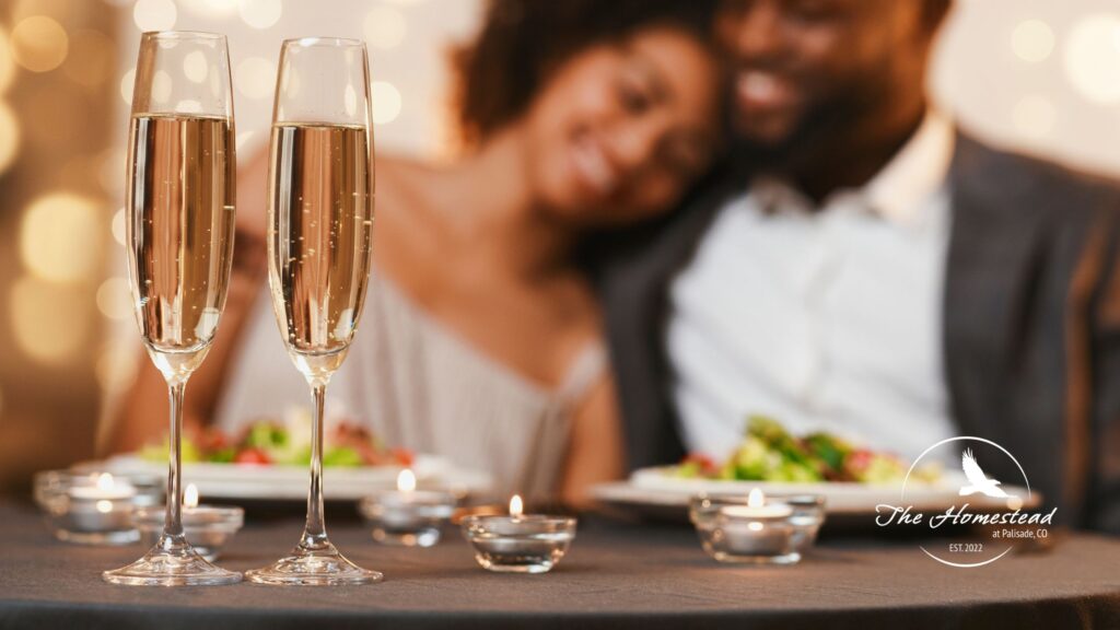 Two champagne glasses and small candles on a table set the mood for a romantic winter escape, while a smiling couple enjoys dinner in the background. The Homestead logo graces the lower right for an inviting touch.