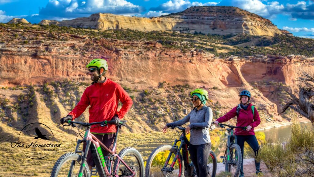 Three cyclists in helmets and jackets stand with their bikes on a desert trail, smiling as they admire scenic red rock cliffs and mesas beneath a blue sky. Experience the Best Spring Hikes and Bike Rides Palisade has to offer. The Homestead logo appears in the lower left corner.