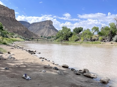 A calm river flows between rocky cliffs and green trees under a bright blue sky with scattered clouds. The riverbank is sandy with scattered rocks, and distant plateaus rise in the background.