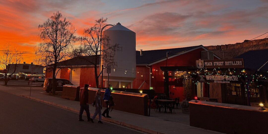 People walk past a distillery and bar with string lights at sunset. The sky is vibrant orange and pink, silhouetting trees and buildings on a quiet street.